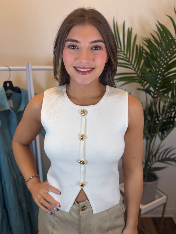 Woman wearing sleeveless white top with round neck and gold sun and seashell details in clothing store setting.