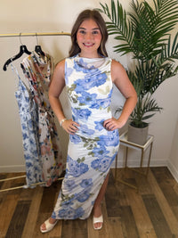 Woman wearing a sleeveless floral maxi dress standing in a room with a rack of dresses and a plant.