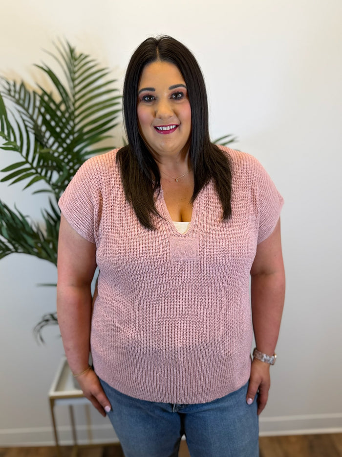 Woman wearing a pink knitted top and blue jeans standing indoors with a plant in the background