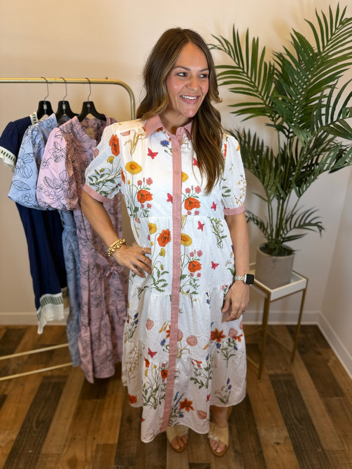 Woman wearing a white floral midi dress with button front detail and pink trim standing in front of a rack of clothes with plants in the background.