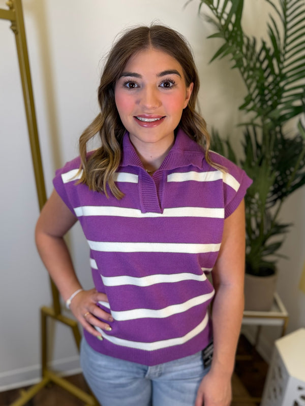 A woman wearing a purple and white horizontally striped knit polo shirt with cap sleeves, paired with light blue jeans, standing in an indoor setting with a gold mirror and a green potted plant in the background.