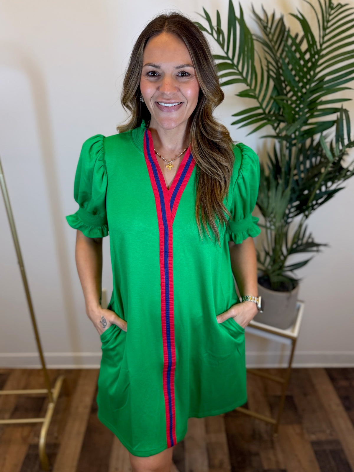 Woman wearing a green dress with red and blue stripe and short puff sleeves indoors with her hands in her pockets.