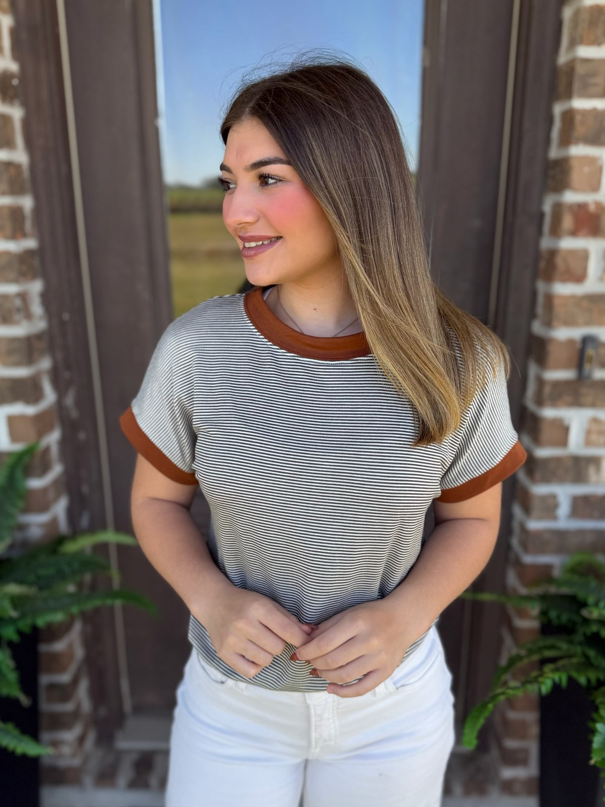 A woman wearing a short-sleeve ringer t-shirt with thin olive and white horizontal stripes and brown trim on the crew neck and sleeve cuffs, paired with white pants, standing in front of a brick door.