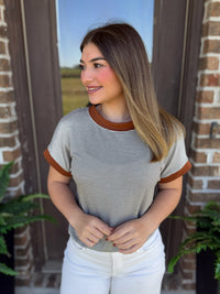 A woman wearing a short-sleeve ringer t-shirt with thin olive and white horizontal stripes and brown trim on the crew neck and sleeve cuffs, paired with white pants, standing in front of a brick door.