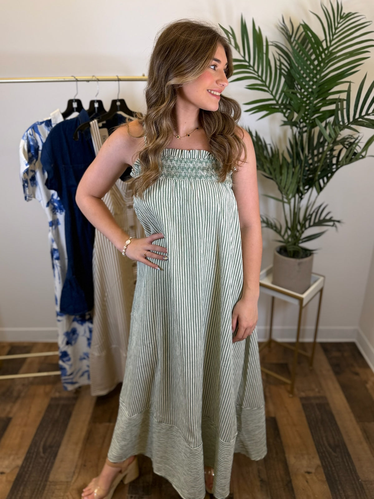 Woman wearing a sleeveless green striped dress in a room with plants and clothing racks.