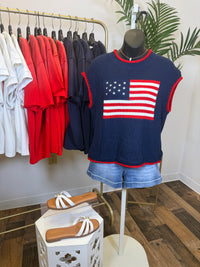 Mannequin wearing a short sleeve navy sweater with an American flag design and red trim, and denim shorts, surrounded by hanging clothes and shoes.
