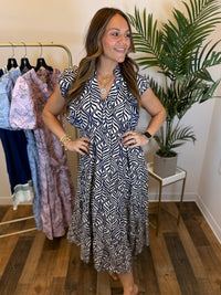Woman in a patterned maxi dress with ruffled sleeves and split neckline standing in a clothing store with racks of clothes and plants in the background.