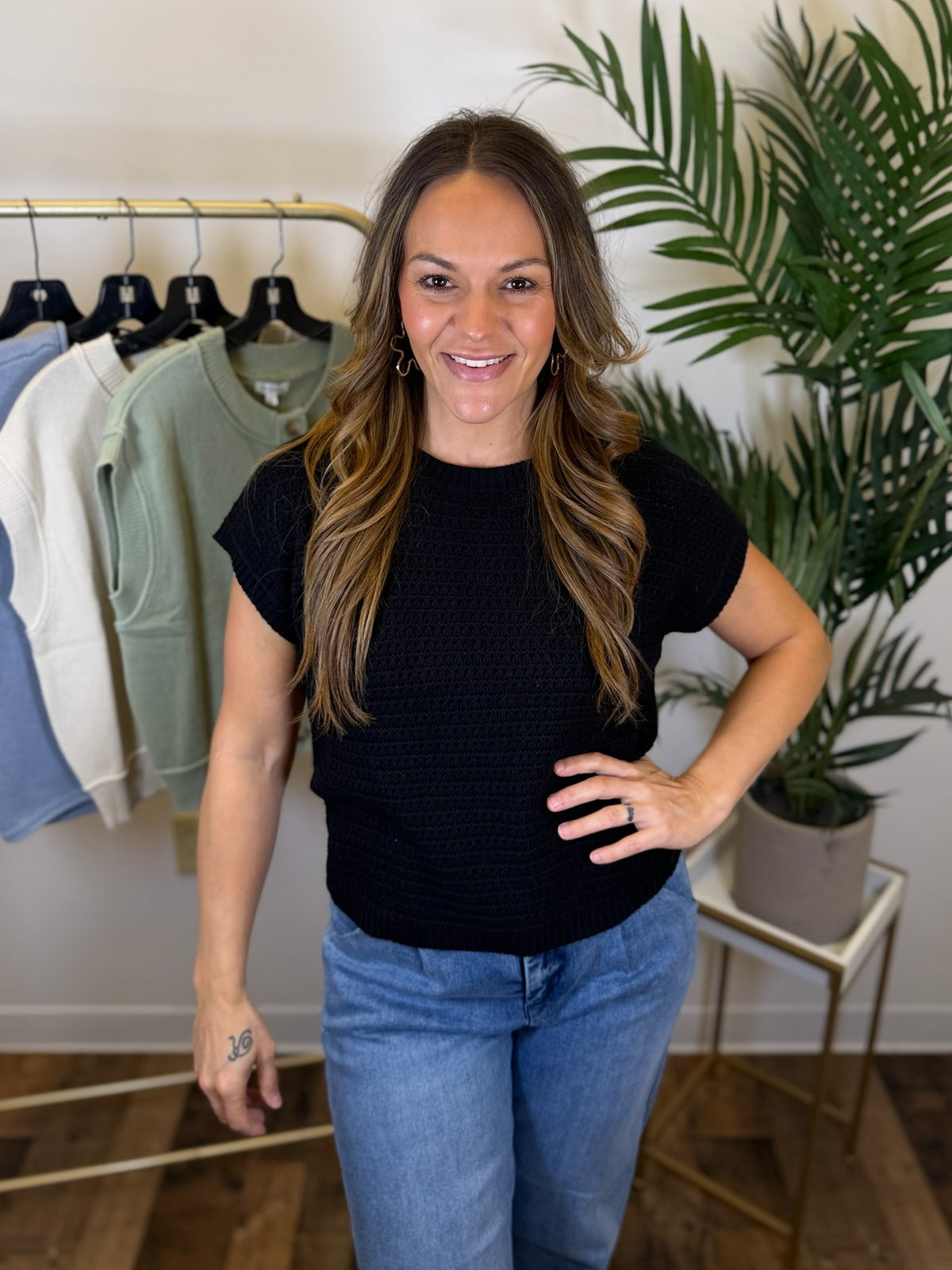 Woman wearing a black short sleeve knit t-shirt and blue jeans standing in a room with clothing racks and plants.
