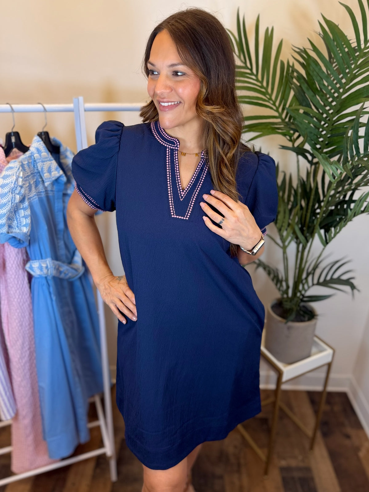 Woman wearing a navy mini dress with puff sleeves and pink trim standing in a room with a clothing rack and plant.