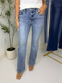 Person wearing medium wash bootcut blue jeans standing in a room with a plant and clothing rack.