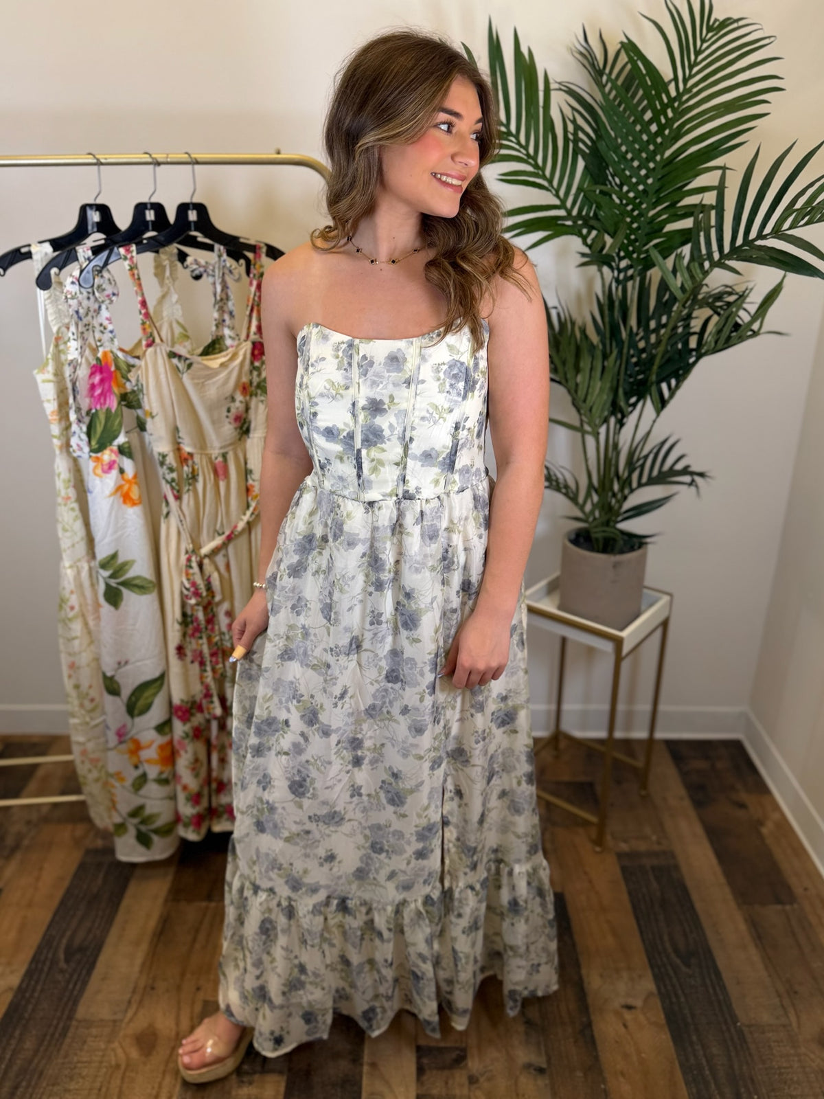 Woman wearing a blue and cream floral midi dress standing in a room with plants and clothing racks.