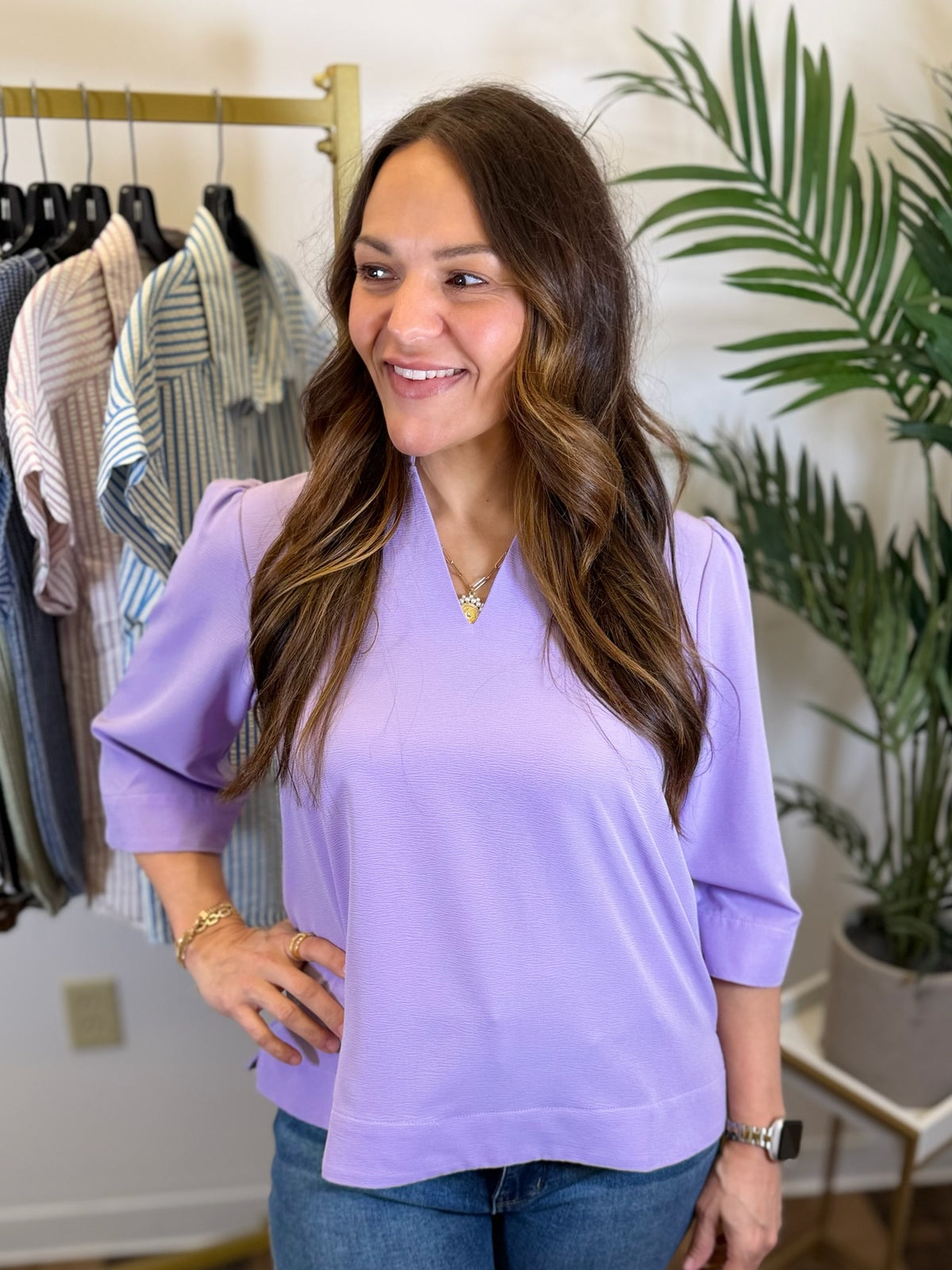 Woman wearing a v neck Lavender blouse with quarter length sleeves in a clothing store with plants in the background