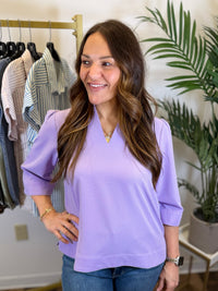 Woman wearing a v neck Lavender blouse with quarter length sleeves in a clothing store with plants in the background