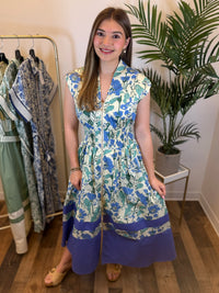 Woman wearing a sleeveless floral midi dress with v neck and zip front standing in a room with plants and clothing racks.