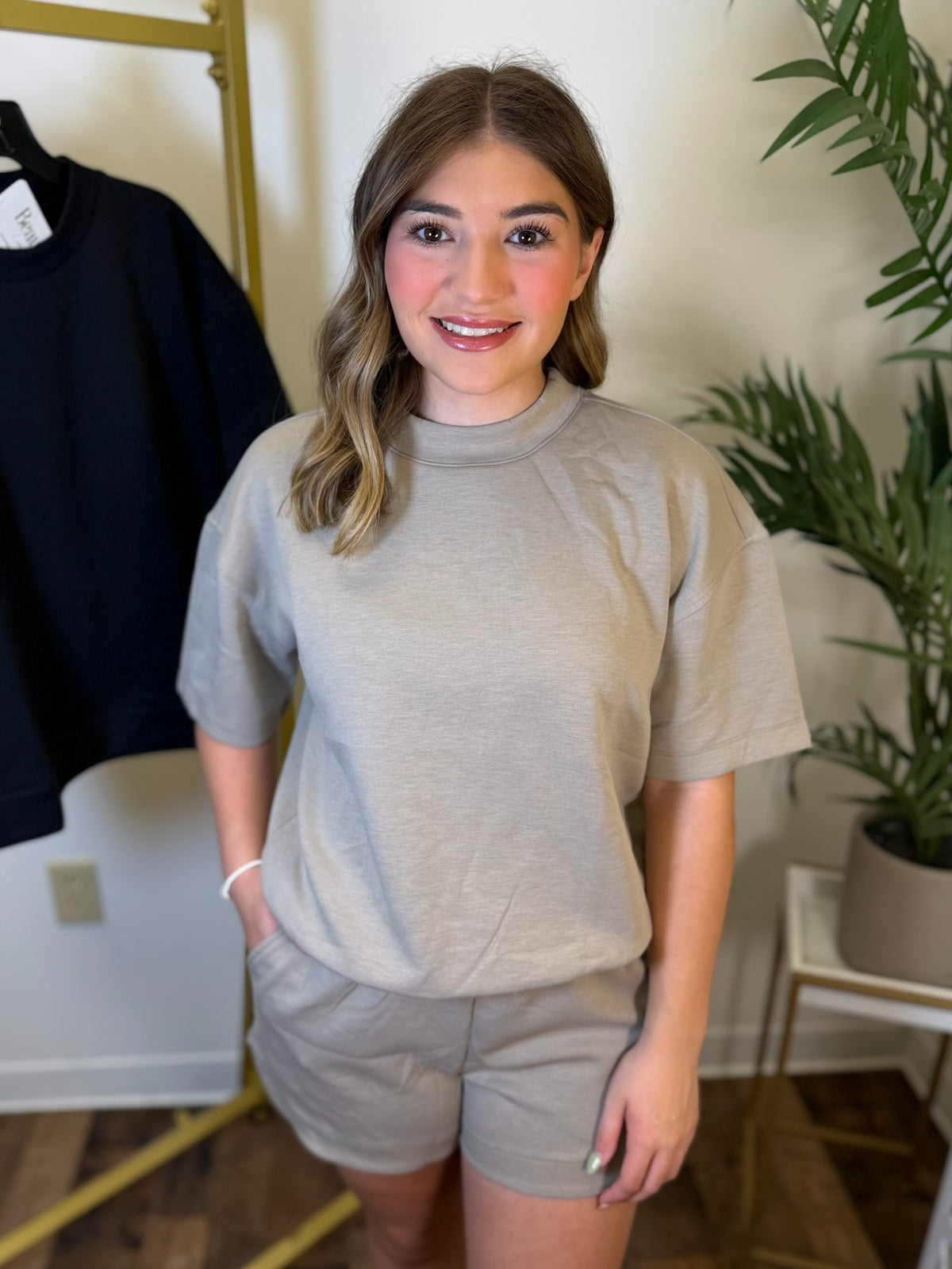 A woman modeling a light beige short-sleeve mock neck t-shirt and matching shorts set, standing in a room with a gold clothing rack and a potted plant.
