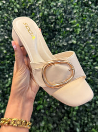 Beige heeled sandal with gold buckle held by a hand against a green hedge background