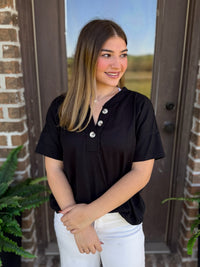 A woman wearing a black short-sleeve henley-style top with three buttons and white pants, standing in front of a brown wooden door with brick columns.