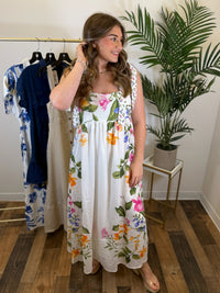 Woman wearing a floral maxi dress with tie strap details standing in a room with a clothing rack and plant.