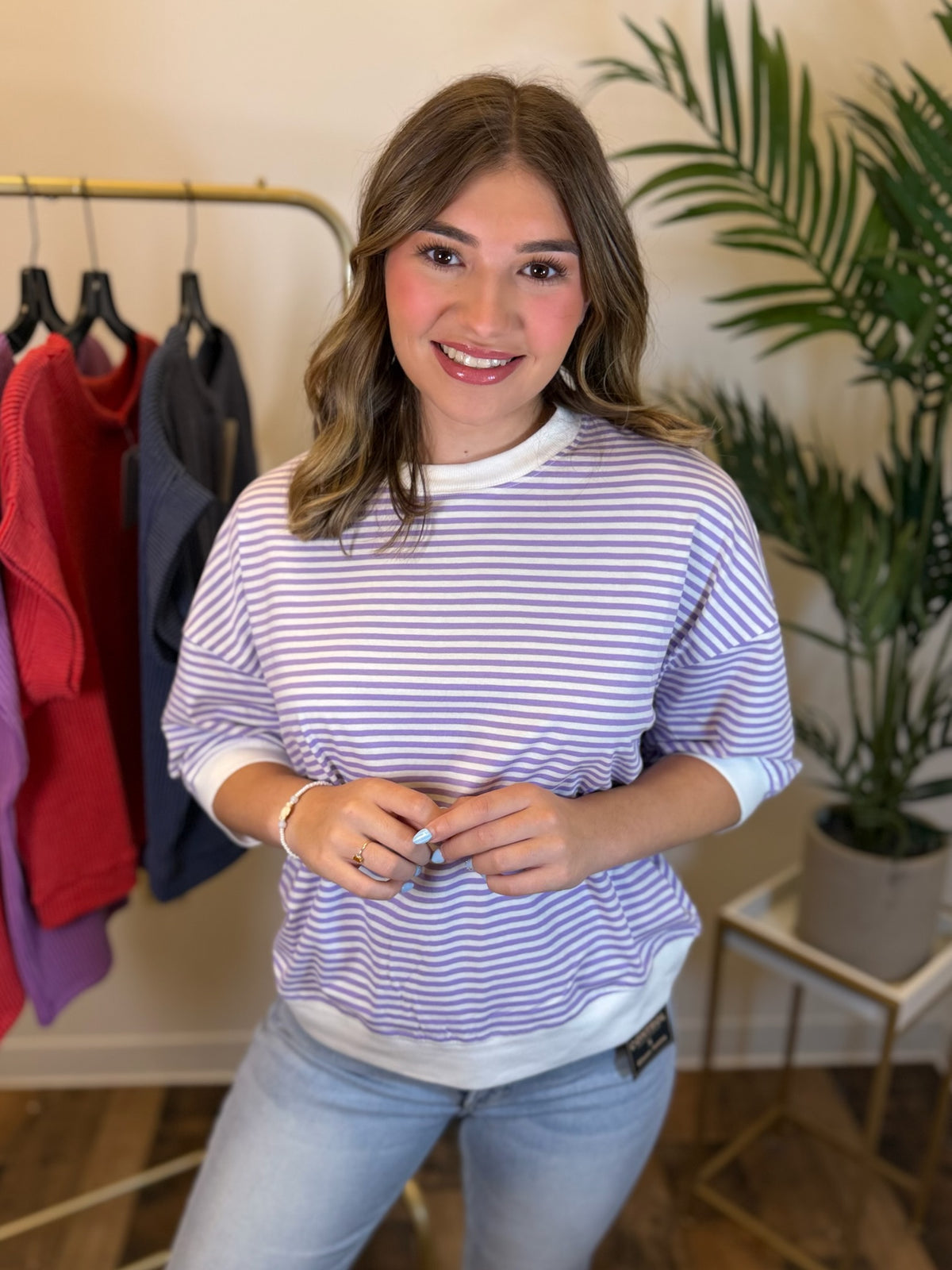 A woman wearing a lavender and white striped short-sleeve t-shirt with white trim, paired with light blue jeans, standing in a boutique setting with a clothing rack and a potted plant in the background.