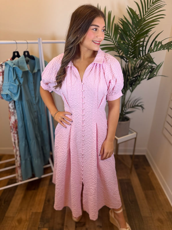 Woman wearing a pink textured dress with a button front in a clothing store with plants and racks of clothes in the background.