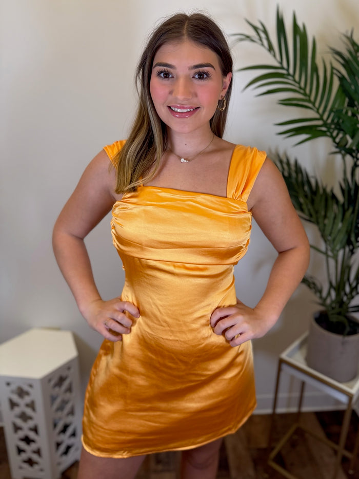 A woman modeling a short, satin-like orange dress with a square neckline and ruched bodice, standing indoors against a light wall with a potted plant in the background.