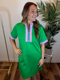 Woman wearing a green dress with pink and blue accents, standing indoors with a plant in the background.