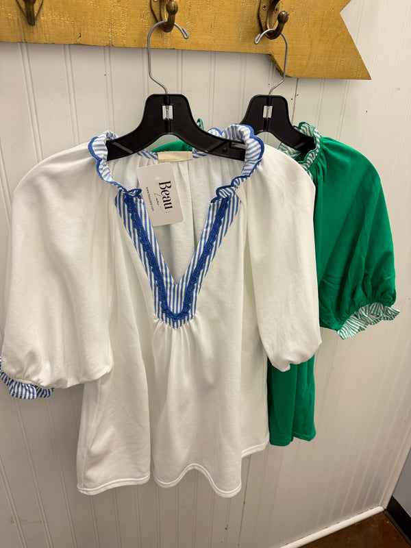 A white short-sleeve blouse with blue striped ruffle trim and a V-neckline, hanging on a black hanger. A green blouse is partially visible behind it on a wooden wall rack.