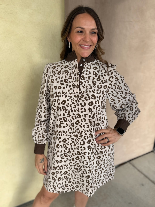 A woman modeling a long-sleeve leopard-print mini dress with brown cuffs, standing against a beige stucco wall.