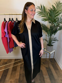 A woman modeling a black short-sleeve maxi dress with a beige center panel and collar, standing in a boutique setting with a clothing rack and a potted plant in the background.