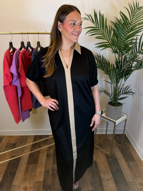A woman modeling a black short-sleeve maxi dress with a beige center panel and collar, standing in a boutique setting with a clothing rack and a potted plant in the background.