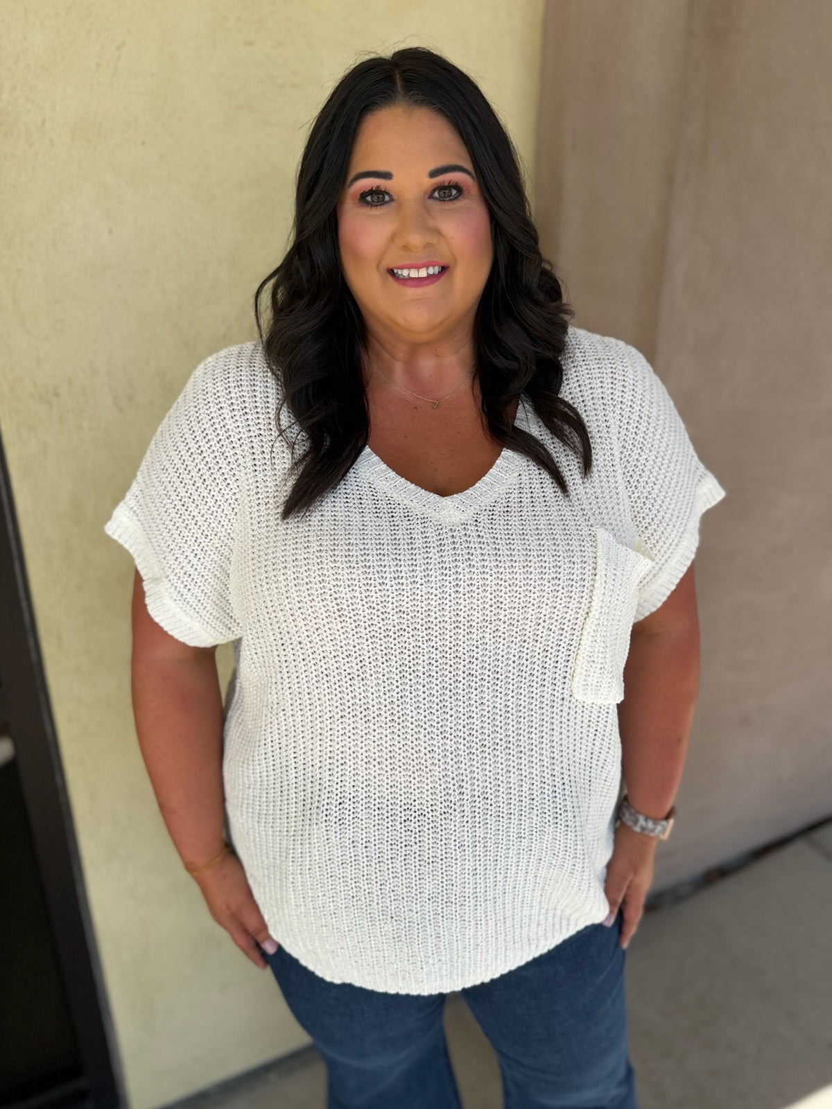 A woman wearing a white textured knit short-sleeve V-neck top with a chest pocket, paired with blue jeans, standing against a beige stucco wall.