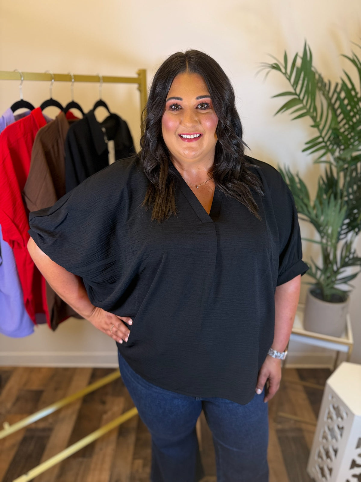 A woman modeling a black short-sleeve blouse with a V-neck and textured fabric, paired with dark blue jeans, standing in a boutique setting with a clothing rack and potted plant in the background.