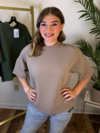 A woman wearing a taupe short-sleeve mock neck top and light blue jeans, standing in a boutique setting with a clothing rack and a potted plant in the background.