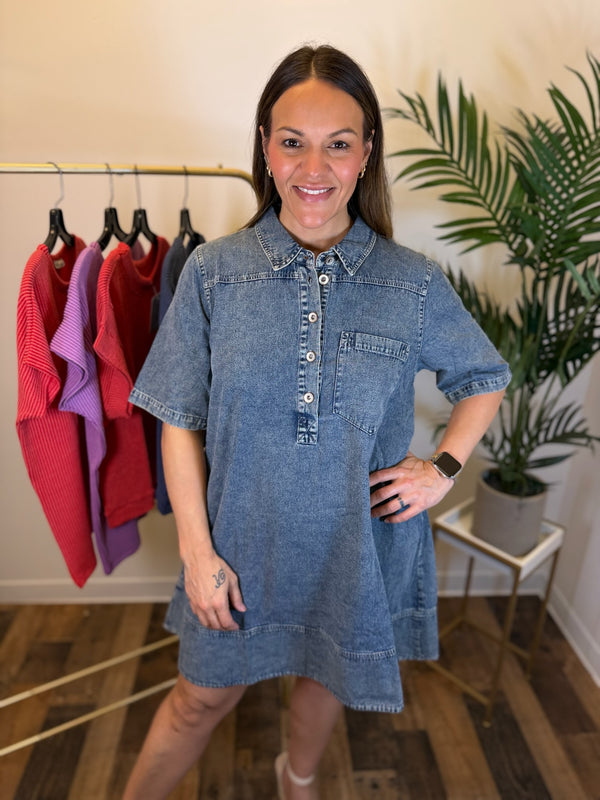 A woman modeling a short-sleeve denim shirt dress in a boutique setting, with a clothing rack and a potted plant visible in the background.