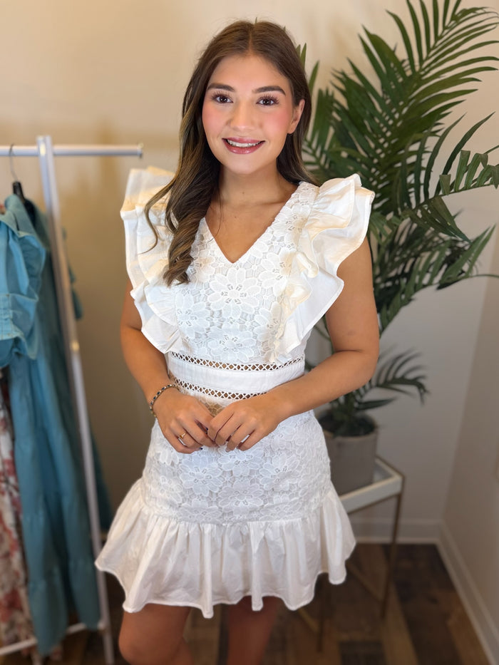 Woman wearing a white lace dress with ruffle sleeve and v neckline standing in a room with plants and clothing racks.