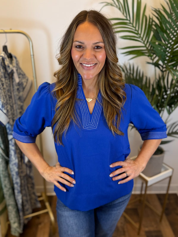 Woman wearing a royal blue blouse with split neckline and stitched trim details in a clothing store setting
