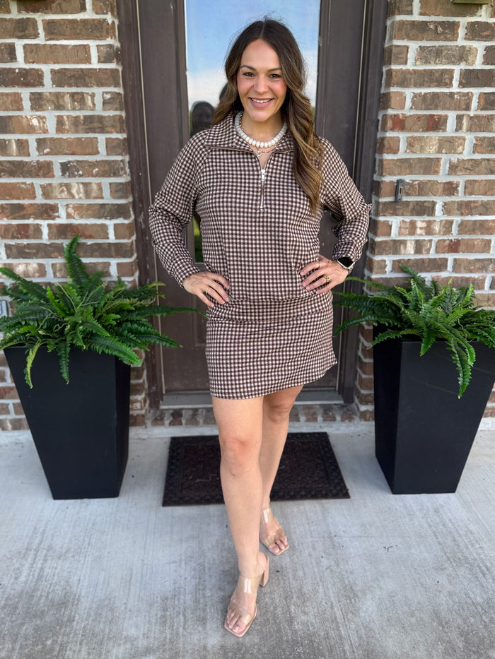 A woman modeling a brown and white houndstooth matching skort and shirt with a half-zip collar, standing on a concrete porch in front of a brick door with potted ferns.
