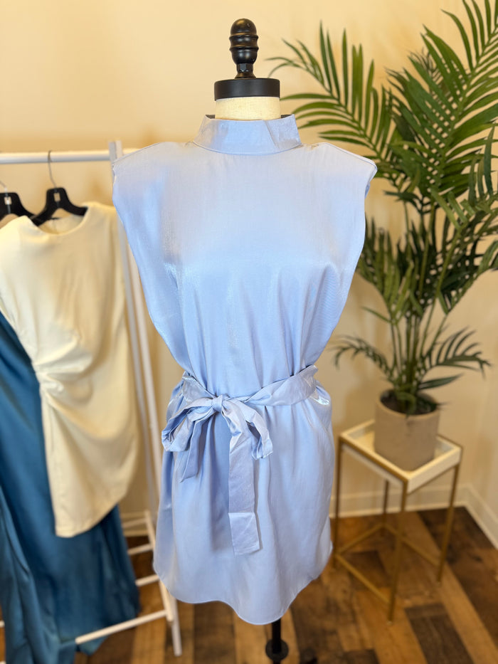 Light blue sleeveless dress with mock neck and waist tie on a mannequin in a clothing store setting with plants and other dresses in the background.
