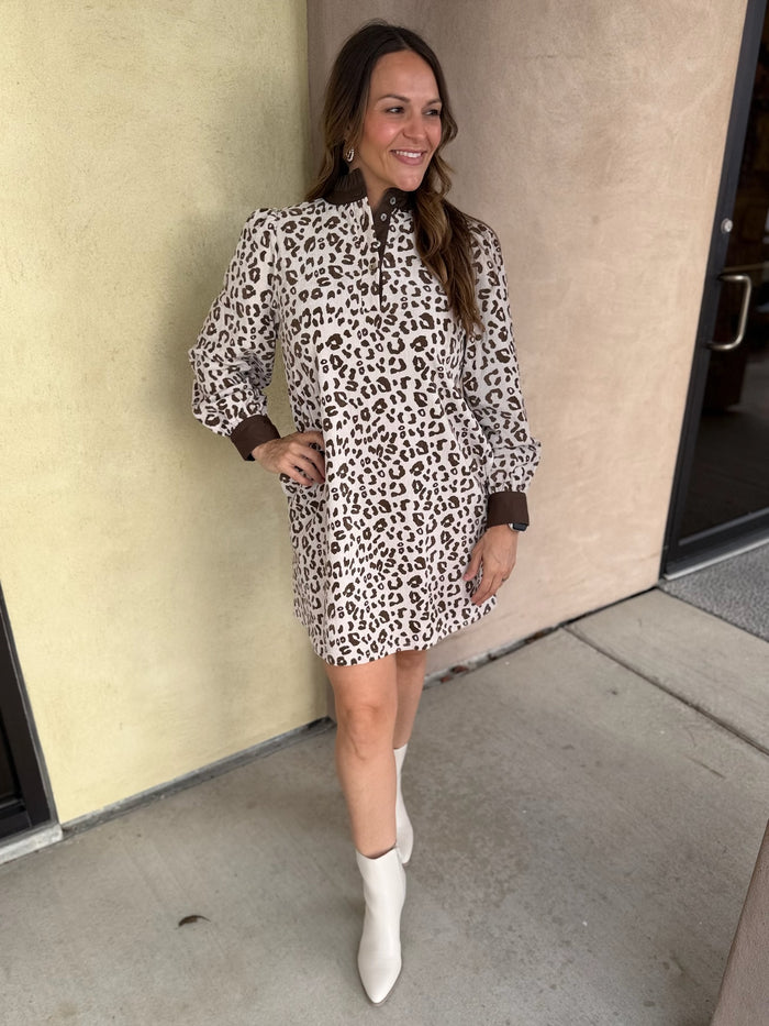 A woman wearing a leopard print mini dress with long sleeves, a ruffled neckline, and brown contrast trim, paired with cream ankle boots.
