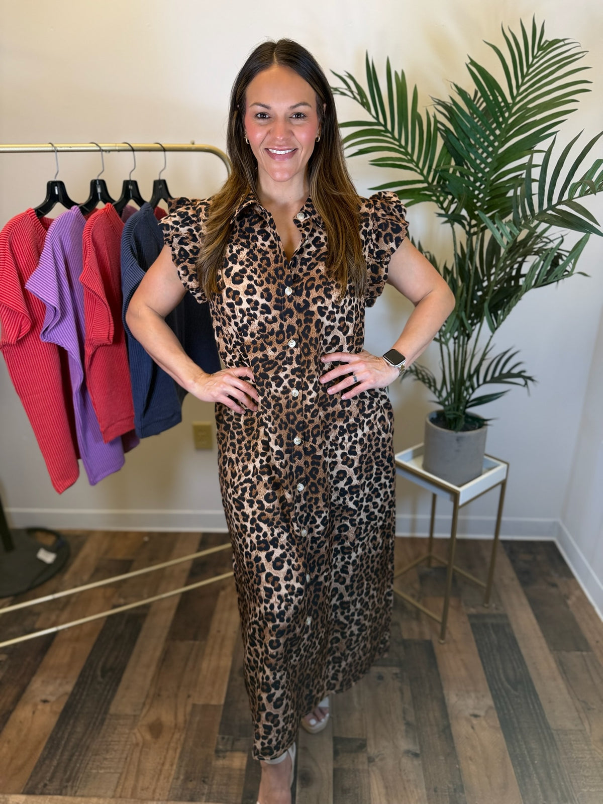 A woman modeling a leopard-print button front midi dress with short ruffled sleeves in a boutique setting, standing in front of a clothing rack and a potted plant.