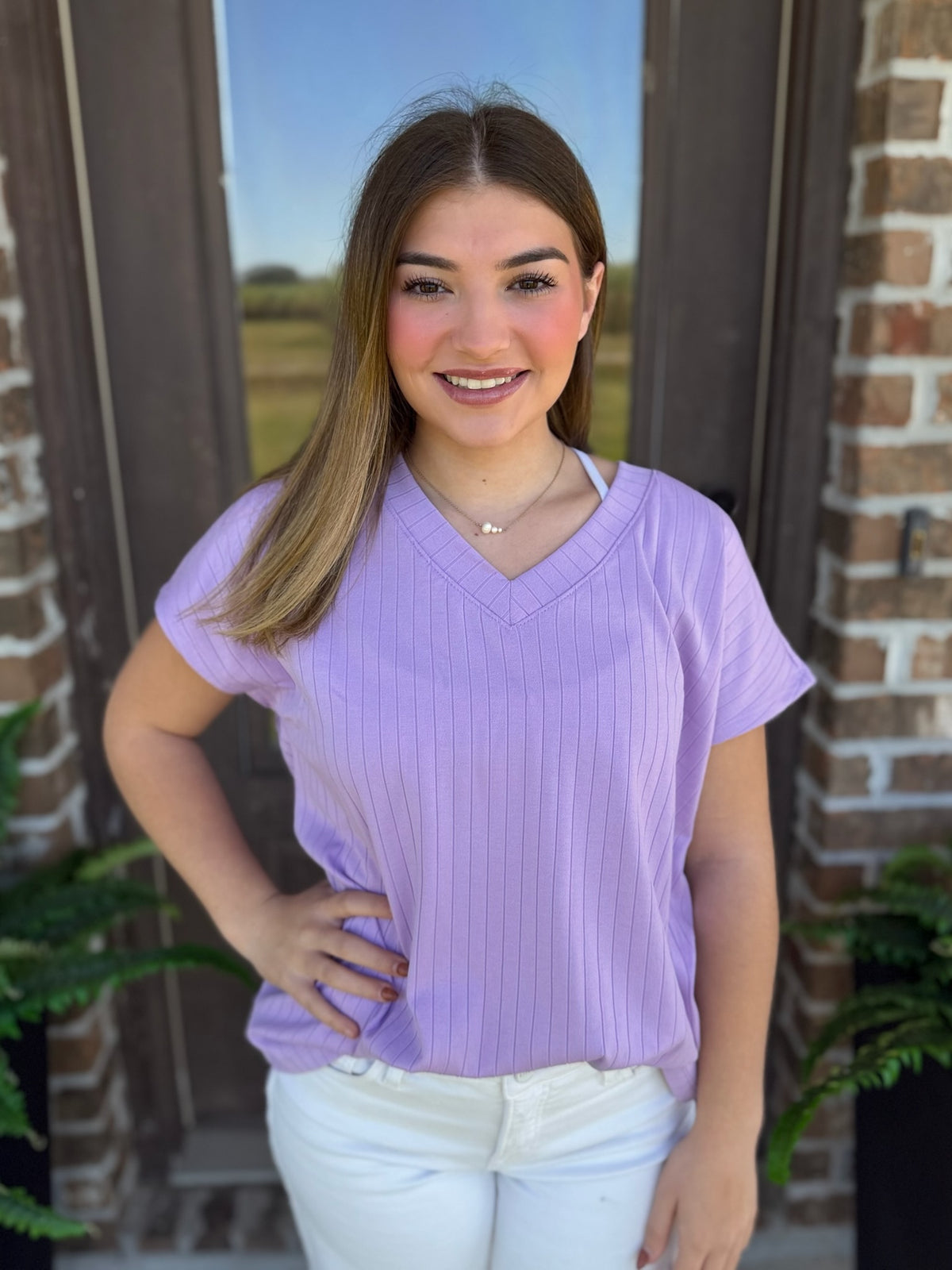 A woman wearing a ribbed lavender V-neck top with short sleeves stands in front of a brick wall.