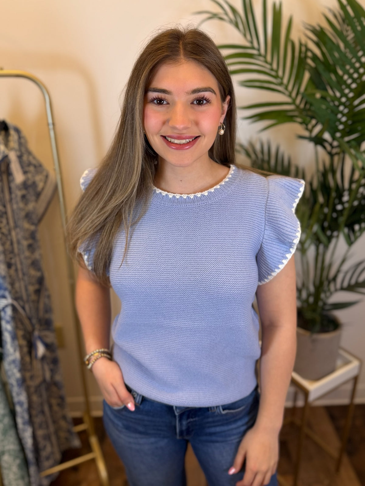 Woman wearing a pale blue knit top with white trim and ruffled sleeves in a room with plants and a rack of clothes.