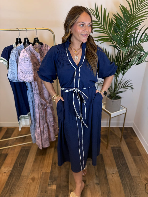 Woman in a textured navy dress with contrast trim details and puff sleeves standing in a clothing store with racks of dresses in the background.