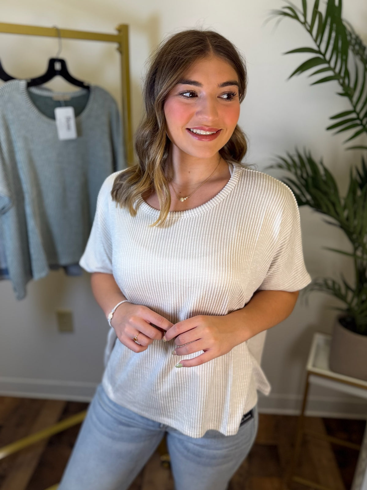 A woman wearing a cream ribbed short-sleeve top and light blue jeans, standing in a boutique setting with a clothing rack and a plant in the background.