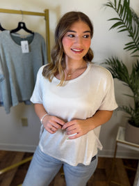 A woman wearing a cream ribbed short-sleeve top and light blue jeans, standing in a boutique setting with a clothing rack and a plant in the background.