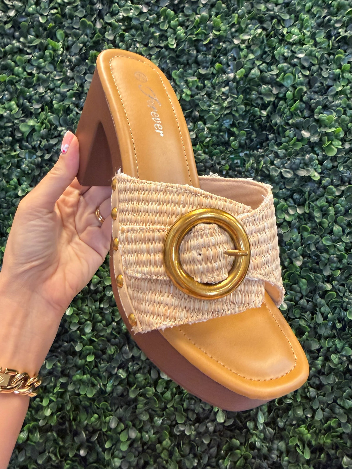 Hand holding a block heel with a natural strap and gold buckle against a green grass background