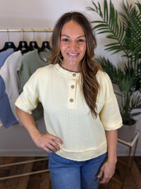 Woman wearing a light yellow short sleeve knit top with a blurred background of a clothing store.