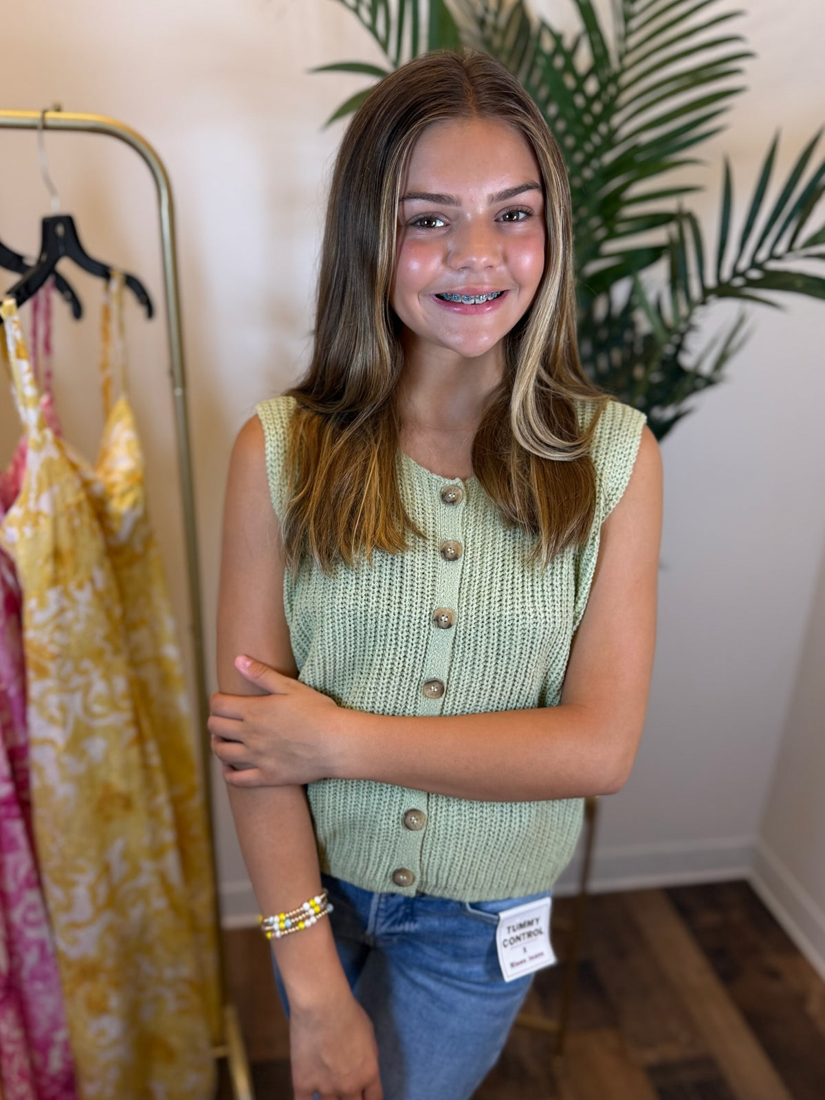 Young girl wearing a sage green sleeveless knit top with buttons, standing in a clothing store.