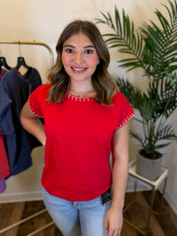 A woman wearing a bright red short-sleeve top with white stitch trim along the neckline and sleeve cuffs, paired with light blue jeans, standing in an indoor boutique setting with a clothing rack and potted plant in the background.
