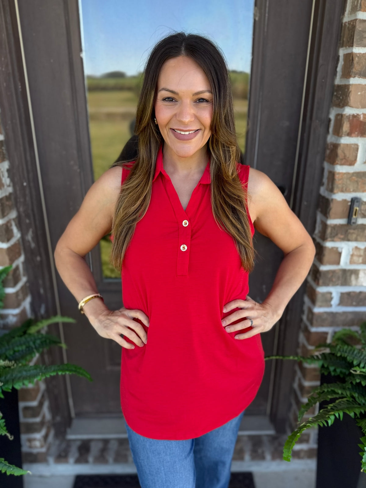 A woman wearing a bright red sleeveless polo shirt with a button placket and blue jeans, standing in front of a brick door with plants visible on either side.